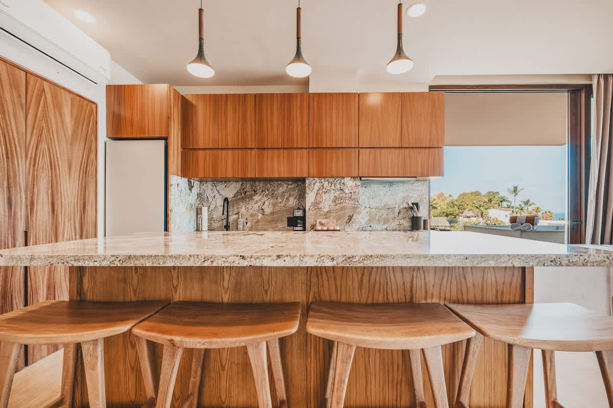 Modern kitchen featuring a large island with granite countertops and four wooden bar stools, rich wooden cabinetry, stainless steel appliances, and pendant lighting, with an ocean view visible through the large windows.