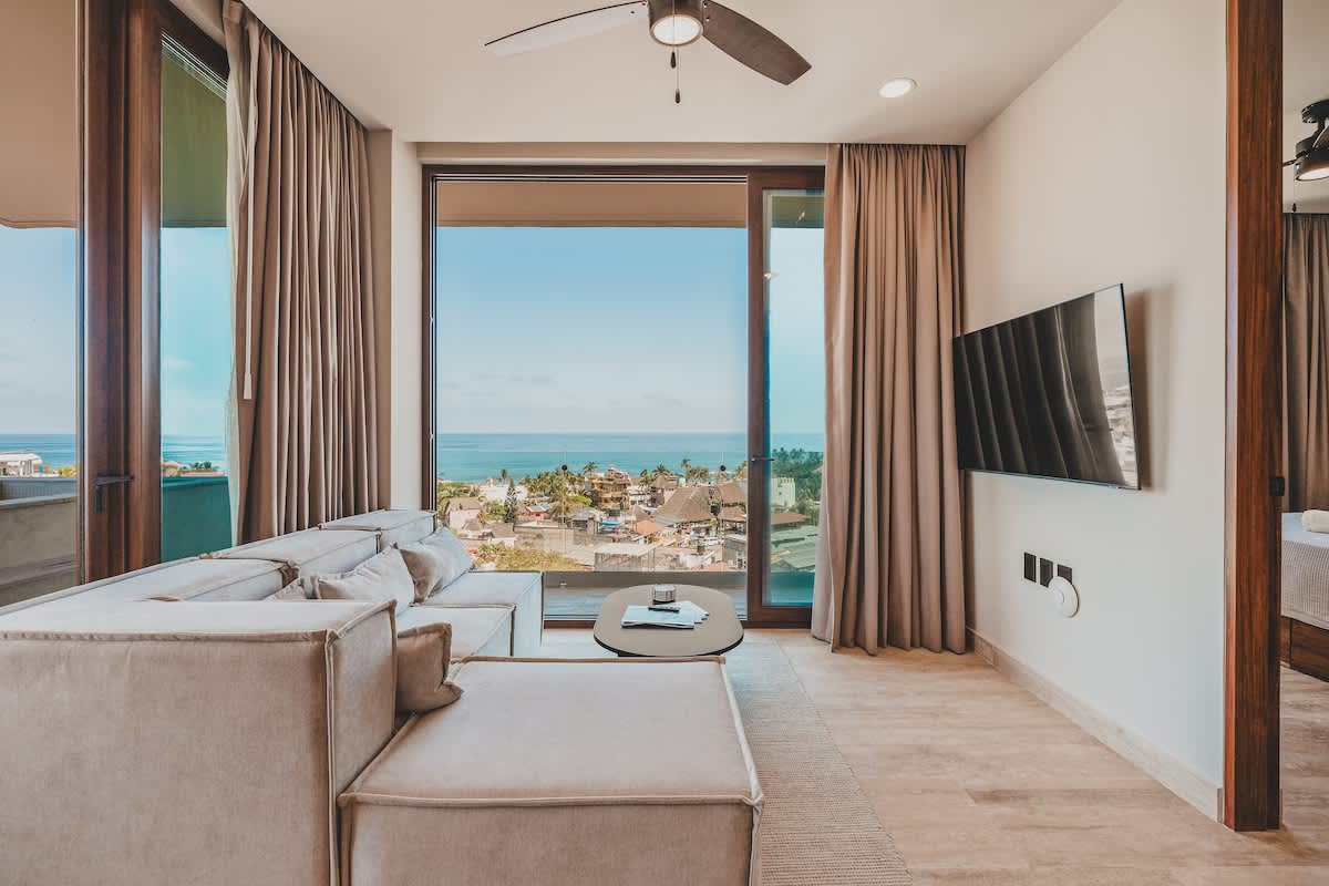 A modern living room features a large beige sectional sofa, a round coffee table, and a wall-mounted TV, with expansive floor-to-ceiling windows providing a stunning ocean and town view.