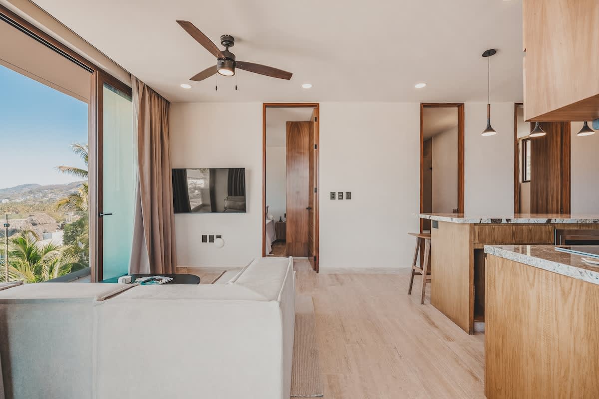 Open-concept living space with a light-colored sectional sofa, wall-mounted TV, and a ceiling fan, seamlessly connecting to the kitchen area with an island.