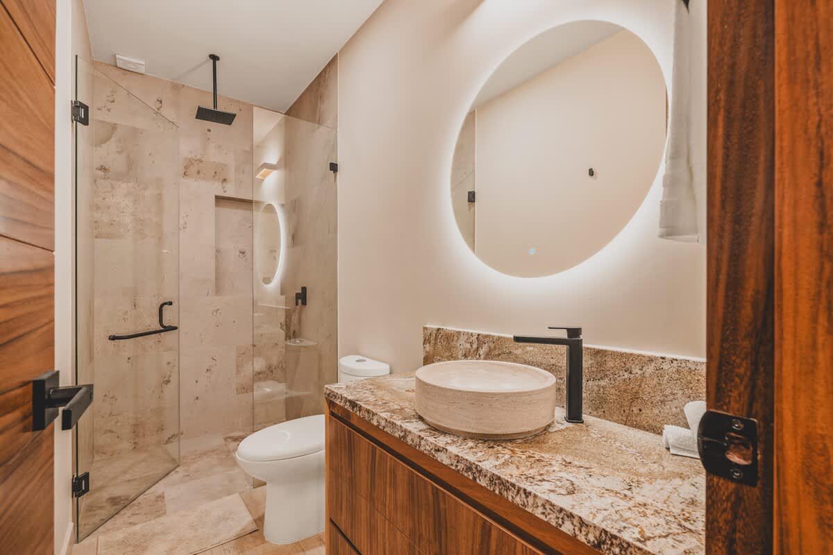 Modern bathroom featuring a glass-enclosed shower with stone-tiled walls, a circular backlit mirror above a vessel sink, and a dark wood vanity with a granite countertop.