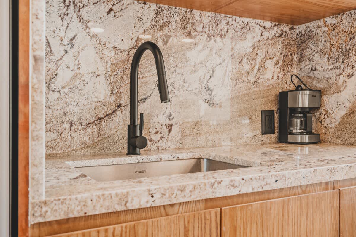 Close-up of a modern kitchen with light granite countertops and backsplash, featuring a black gooseneck faucet over a stainless steel sink, and a coffee maker in the background.