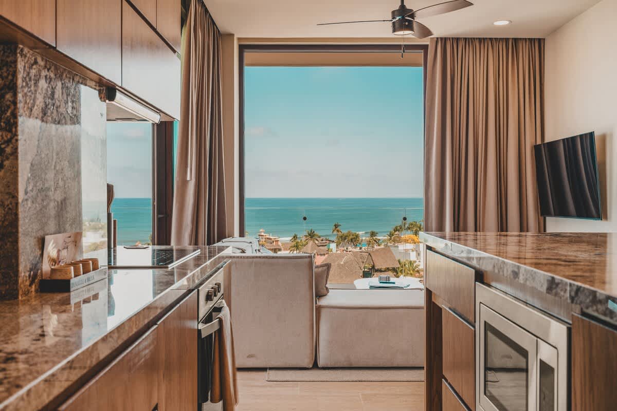 A modern kitchen features dark granite countertops and light wooden cabinetry, seamlessly connecting to the living area with a stunning ocean view visible through large windows.