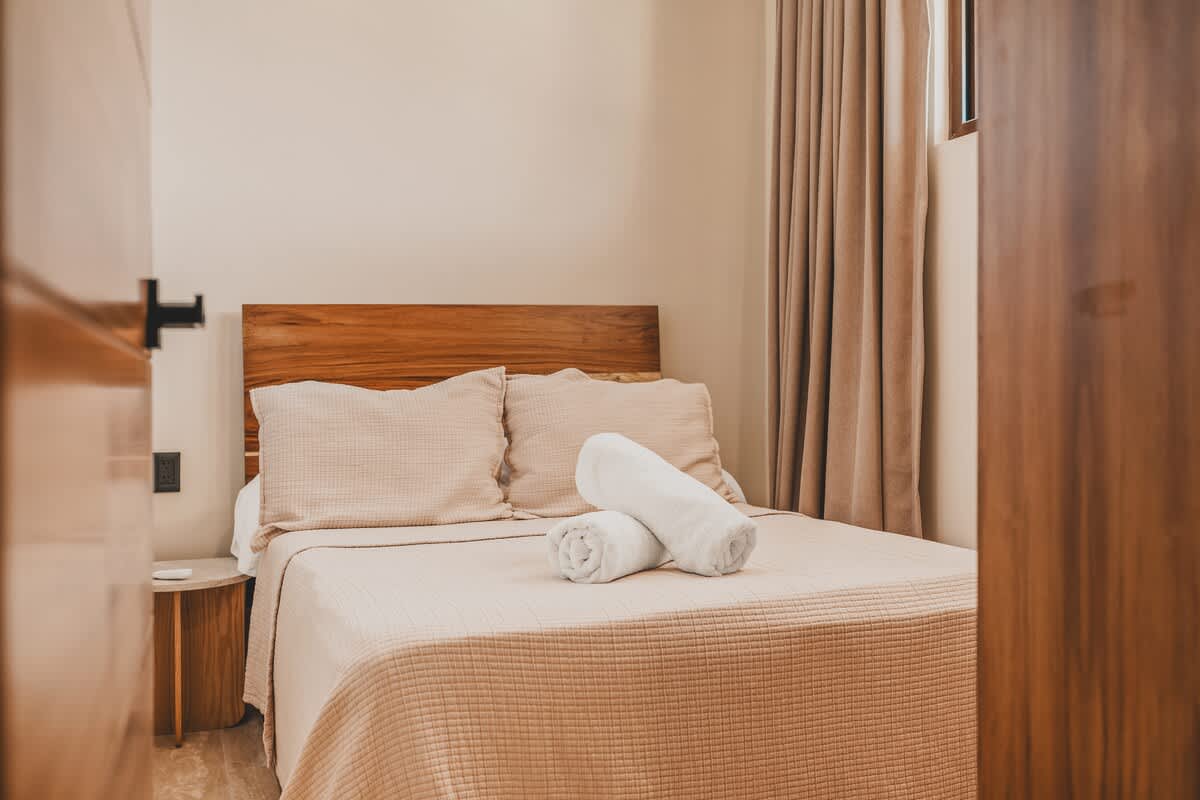 Cozy bedroom with a wooden headboard, light-colored bedding, and two rolled white towels on the bed. A small round side table and light-colored curtains are also visible.
