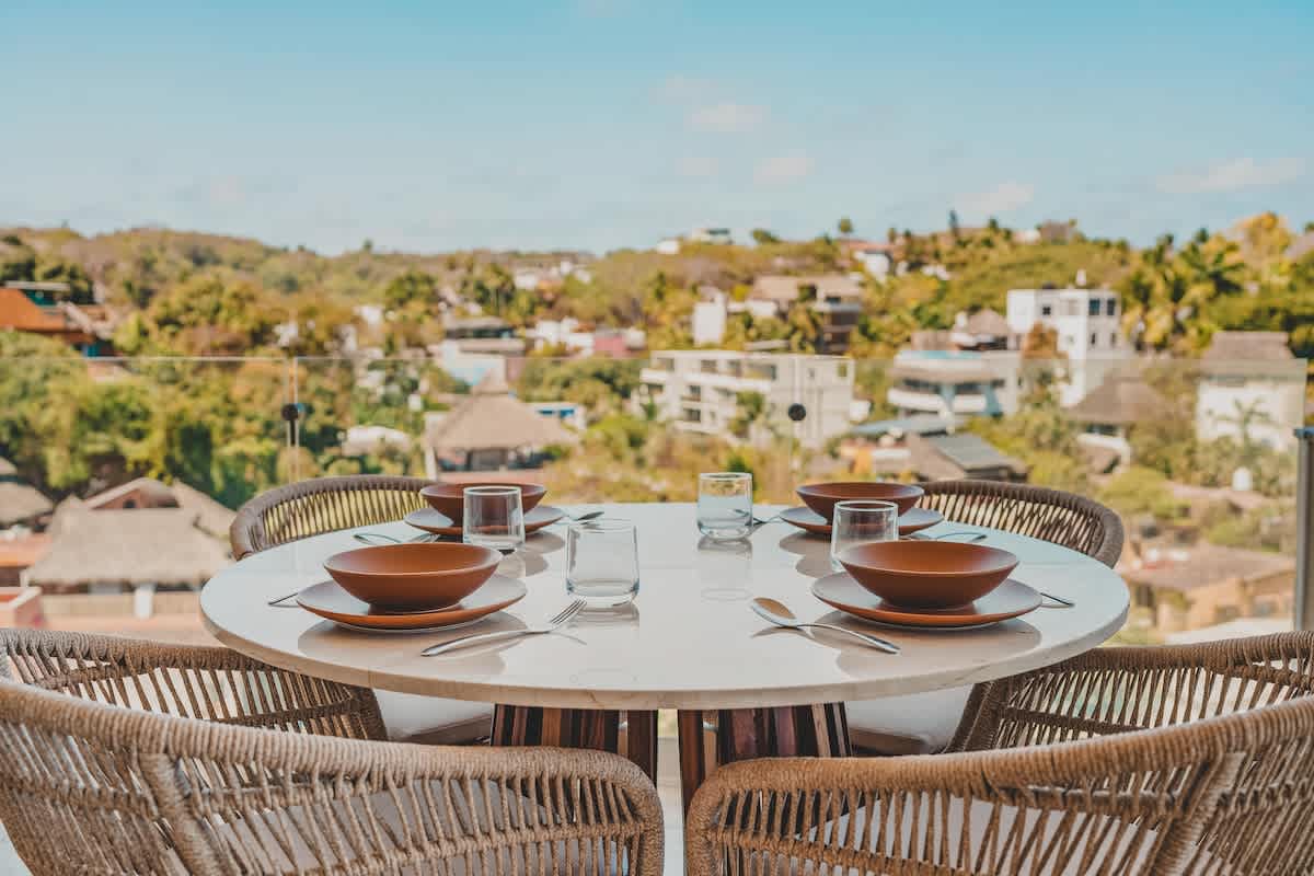 An outdoor dining table, set with rustic bowls and glasses, is ready for a meal on a balcony, offering a scenic backdrop of a green hillside and a charming town.