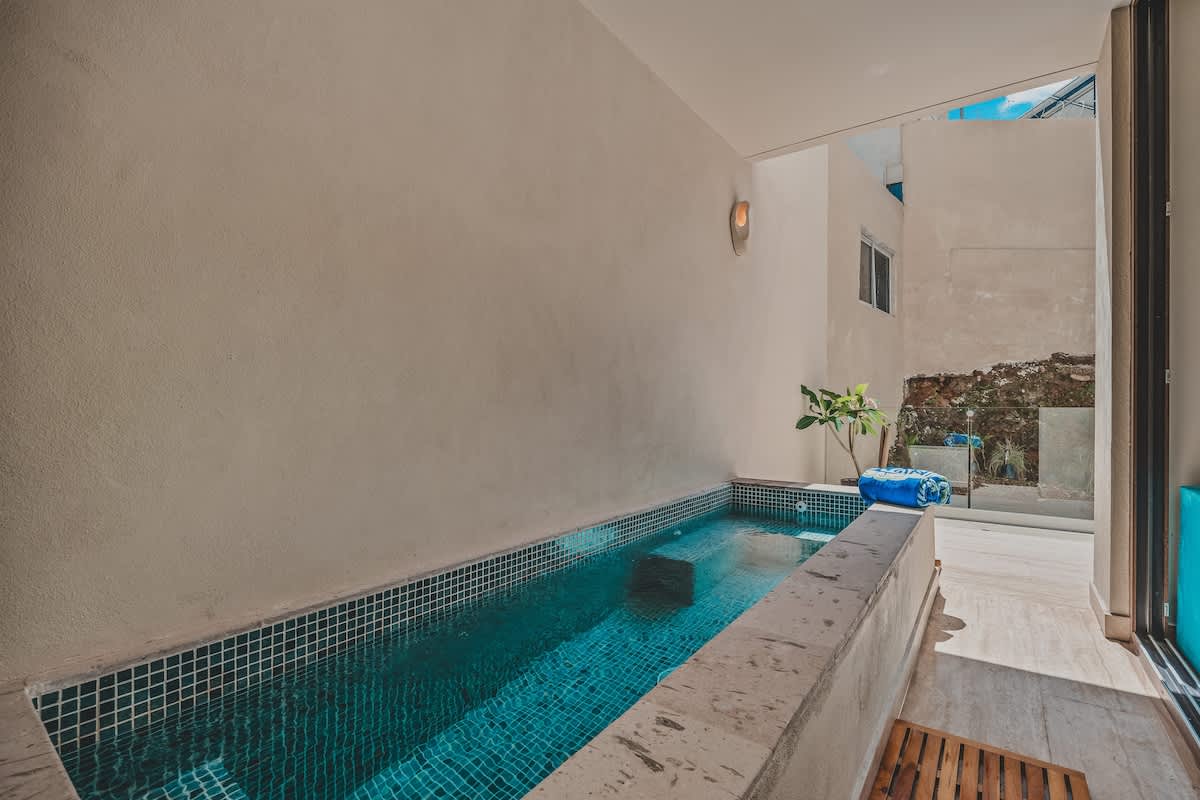 A narrow, rectangular plunge pool with dark blue mosaic tiles is set into a concrete surround, flanked by a light-colored wall and an open-air view, suggesting an intimate outdoor space.