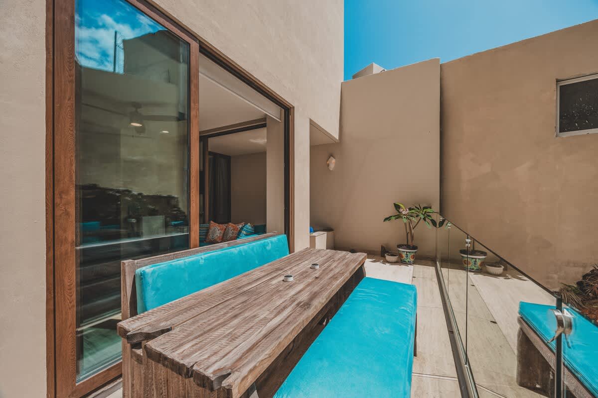 A sunny outdoor patio features a rustic wooden table with built-in benches and bright turquoise cushions, positioned next to a large sliding glass door leading indoors. A glass railing separates the seating area from a small potted plant and light-colored exterior walls.