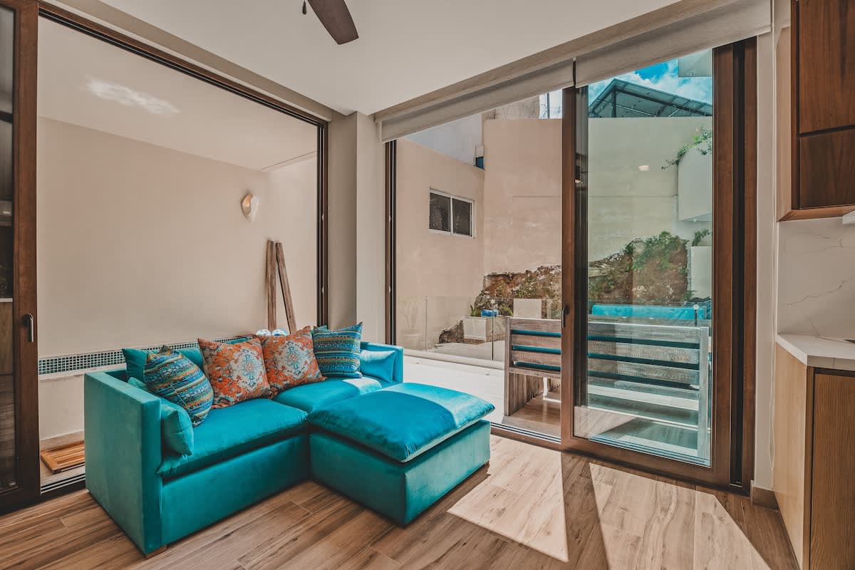 Inviting living room centered around a bright turquoise sectional sofa with decorative pillows, set on light wood-look flooring. Large sliding glass doors open to an outdoor patio area.