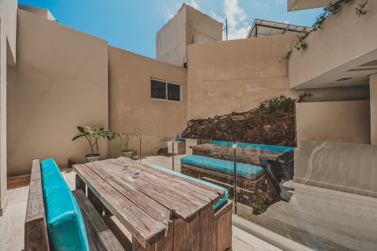 A bright outdoor patio features a rustic wooden table with built-in benches topped with vibrant turquoise cushions, set against light-colored walls and a glass railing. A small potted plant adds a touch of greenery, and a rocky, overgrown area is visible behind another seating arrangement.