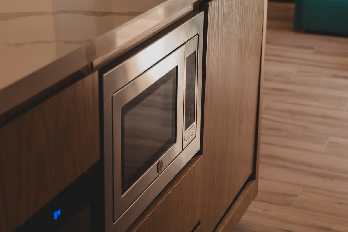 Under-counter built-in stainless steel microwave oven, seamlessly integrated into dark wood kitchen cabinetry, with a light-colored countertop above.