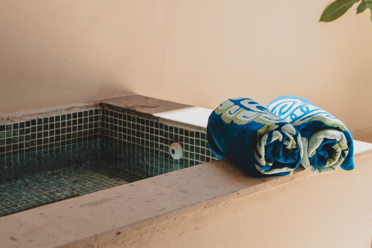 Small, built-in hot tub or plunge pool with blue mosaic tiles, set into a light-colored concrete surround, with two rolled blue and yellow towels.