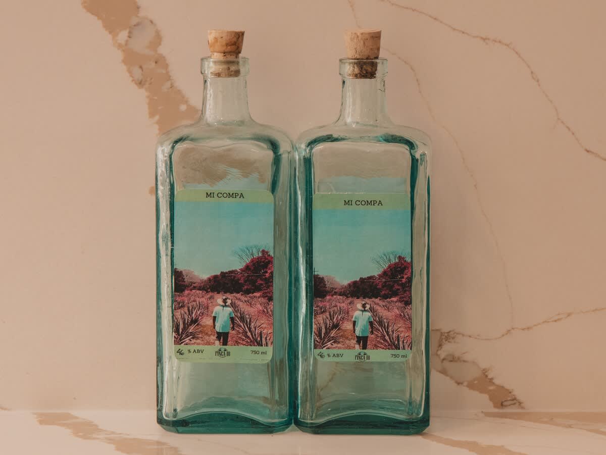 Two identical clear glass bottles with cork stoppers, labeled "MI COMPA" and featuring a scenic desert landscape with a person, stand side-by-side on a light-colored marble-patterned surface. The labels are vibrant with teal and pink hues.