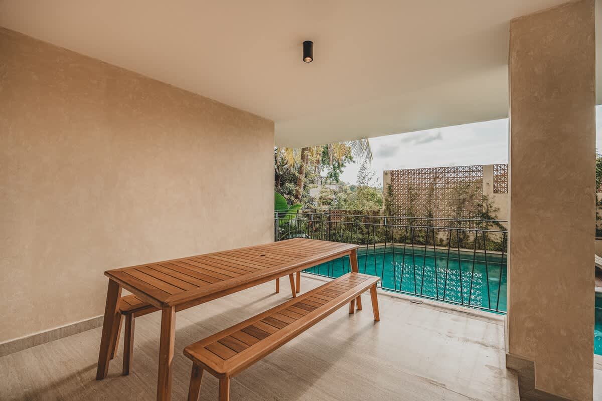 A long wooden picnic table with matching benches is situated on a covered patio, offering a view of a swimming pool and lush green foliage beyond a black metal railing.