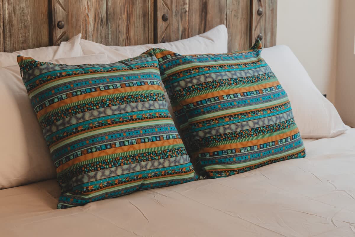 Two vibrant throw pillows with a geometric, tribal-inspired pattern in teal, orange, and green rest on a neatly made bed with white linens. A rustic wooden headboard adds a touch of natural texture to the bedroom decor.