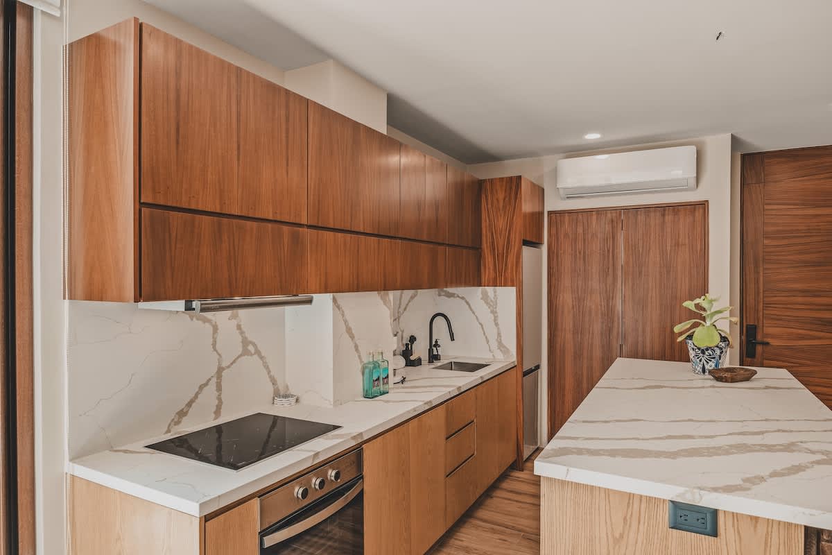 A modern kitchen area showcases a built-in electric stovetop and oven, set within white marble countertops and backsplash, with rich dark wood upper cabinets and lighter wood lower cabinets.