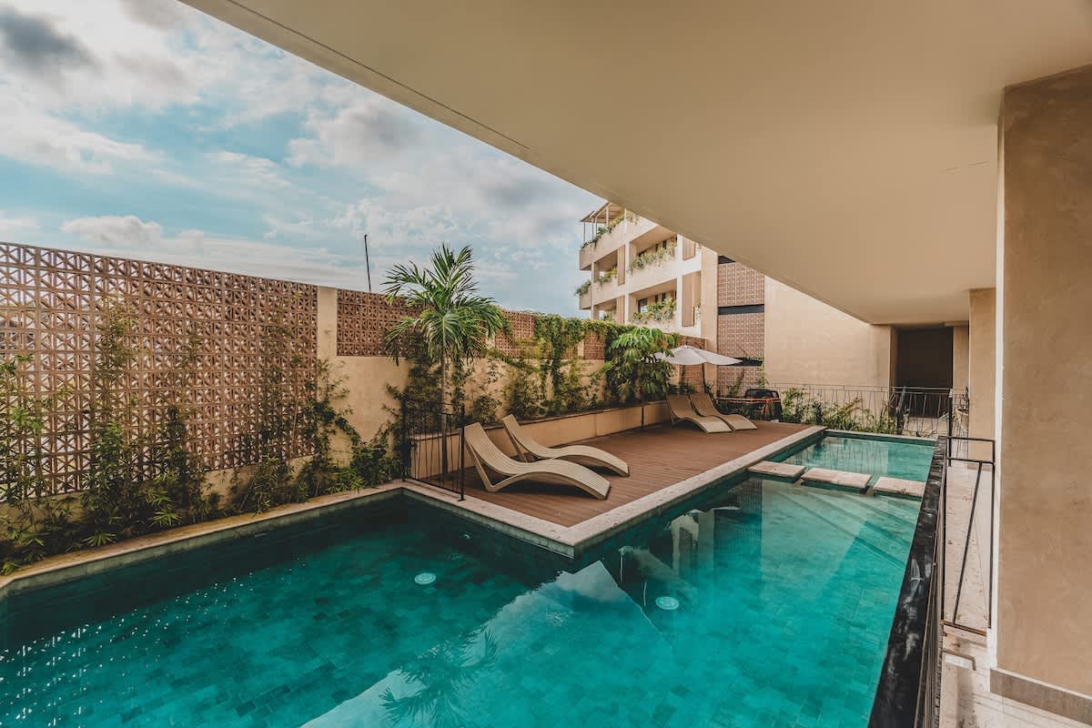 Expansive swimming pool area featuring a long, rectangular pool with a tiled bottom, a wooden deck with multiple lounge chairs, and lush tropical plants against a textured wall.