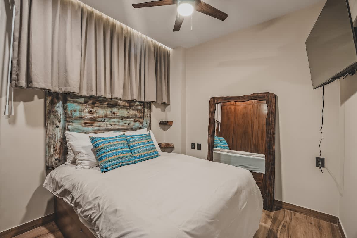 Cozy bedroom with a bed featuring a distressed wooden headboard, light bedding, and two blue patterned pillows. A ceiling fan is visible, along with a large full-length mirror and a wall-mounted TV.