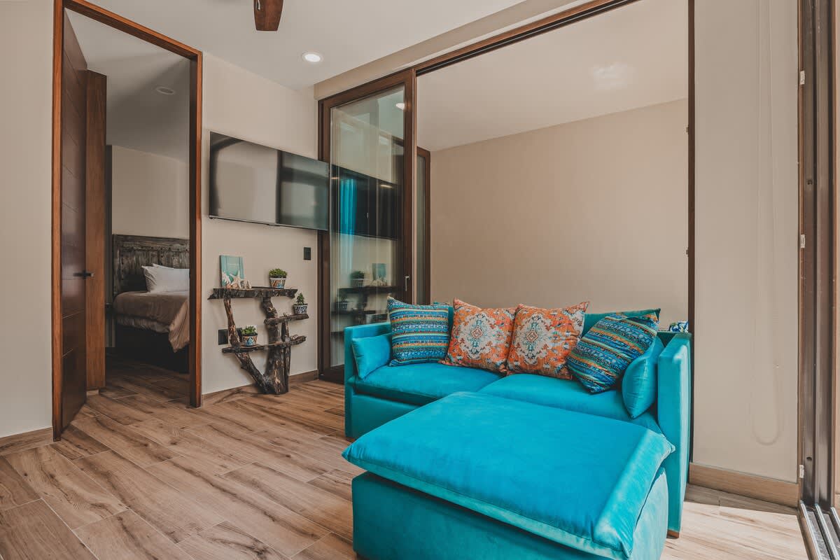 Bright living area with a vibrant turquoise sofa and ottoman, adorned with patterned throw pillows. Wood-look flooring extends throughout, and a wall-mounted TV is visible, with a bedroom entrance nearby.