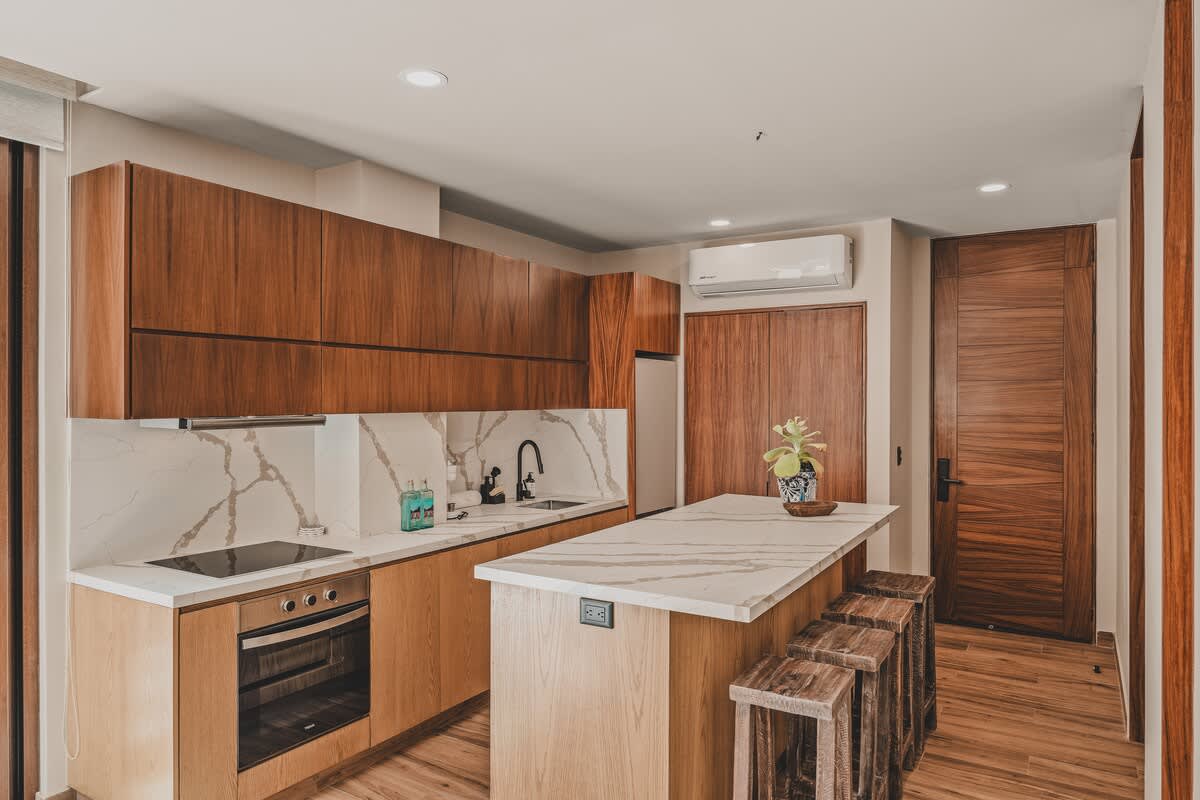 A bright, modern kitchen features a large white marble island with wooden stools, light and dark wood cabinetry, integrated appliances including a refrigerator and oven, and a split air conditioning unit.