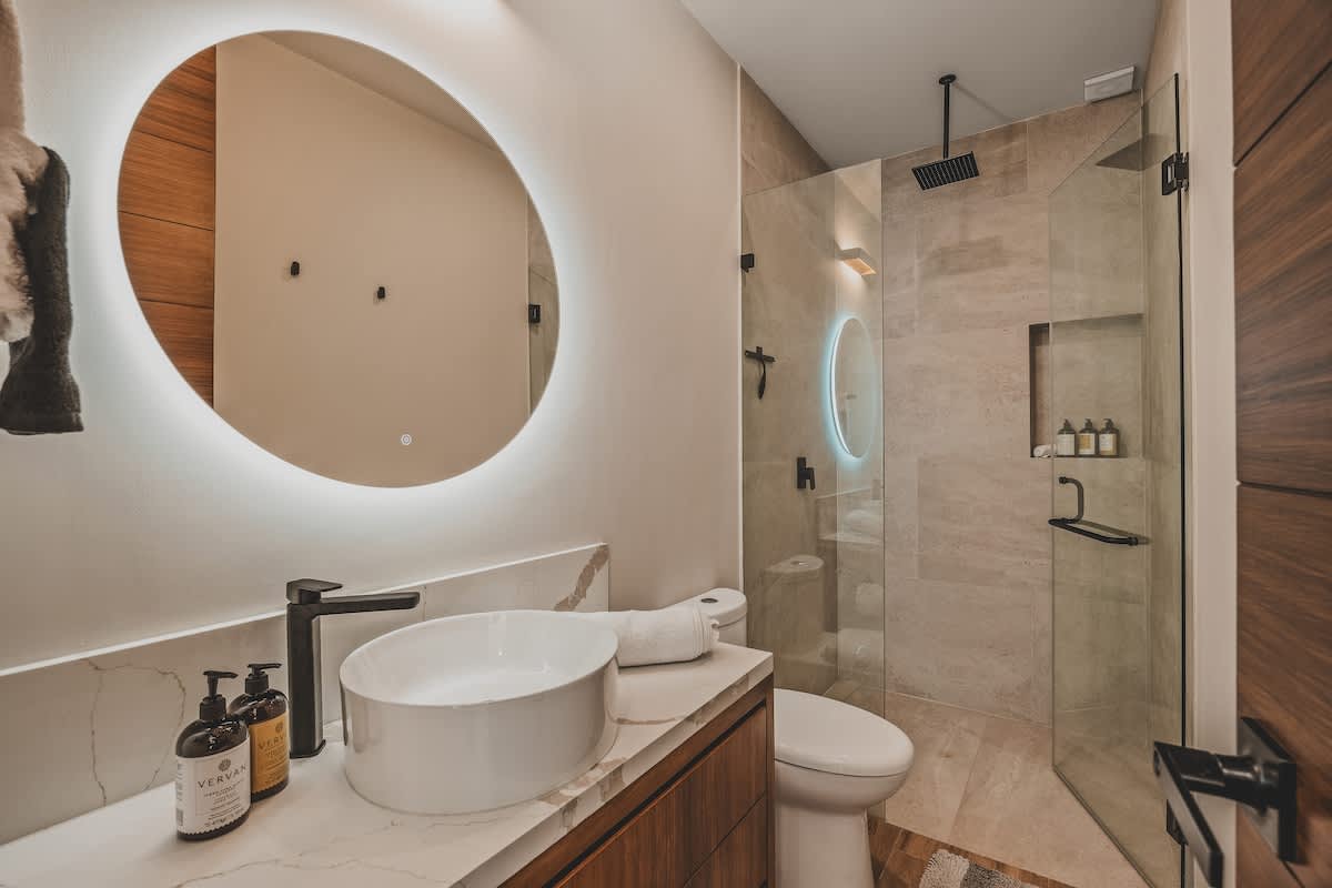 Sleek modern bathroom with a white vessel sink, marble vanity, and a large round backlit mirror. A spacious glass-enclosed shower with a rain showerhead and tiled walls is visible on the right, featuring a shower niche.