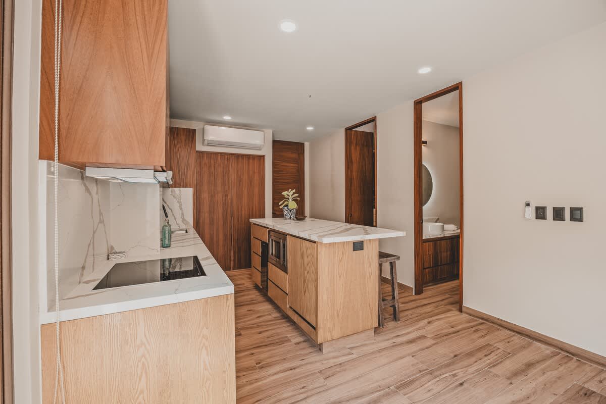 A well-appointed kitchen with light wood cabinetry and white marble countertops extends towards a hallway, revealing a modern bathroom with a circular mirror and dark wood accents.