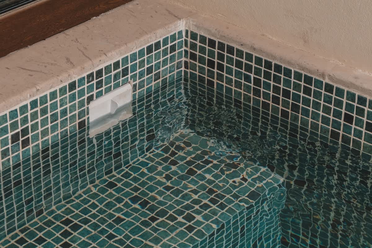 A close-up view of a small, square hot tub or plunge pool, featuring dark teal mosaic tiles with white grout. The water is clear and gently rippling, reflecting the light, with a white skimmer visible on one side.