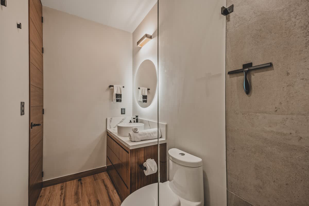 Modern bathroom with a white vessel sink on a marble-topped wooden vanity, a round backlit mirror, and a glass-enclosed shower with a rain showerhead. The floor is wood-look tile.