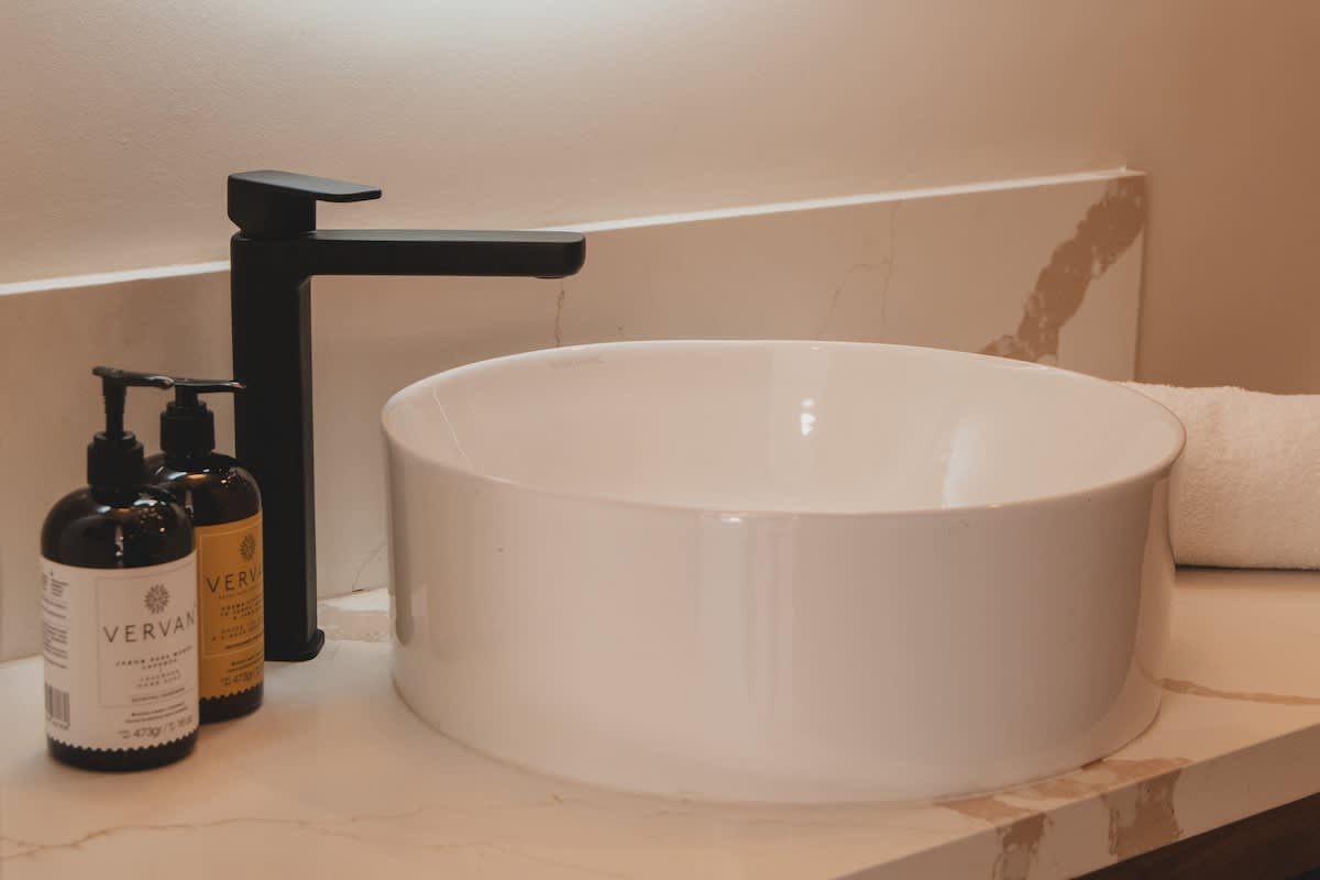 A modern bathroom vanity features a sleek white vessel sink and a matte black faucet, set against a light-colored marble-patterned countertop. Two dark amber bottles of bath products and a white rolled towel are neatly arranged beside the sink.