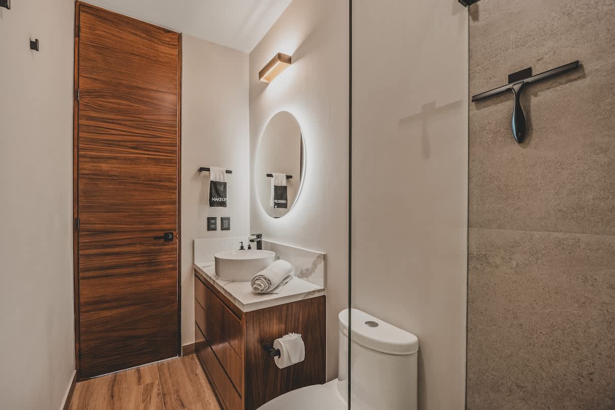 Modern bathroom entrance with a dark wooden door, a white vessel sink on a marble vanity, and a round backlit mirror. A glass panel separates the vanity area from the shower, with a toilet visible.