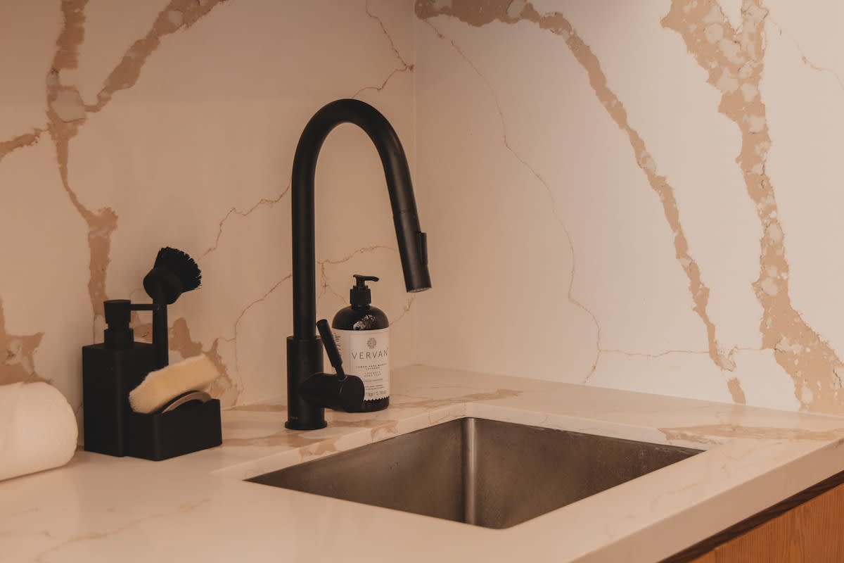 Kitchen sink area with a modern black gooseneck faucet, stainless steel sink, and black soap dispenser and brush holder, against a white and brown marble-patterned backsplash.