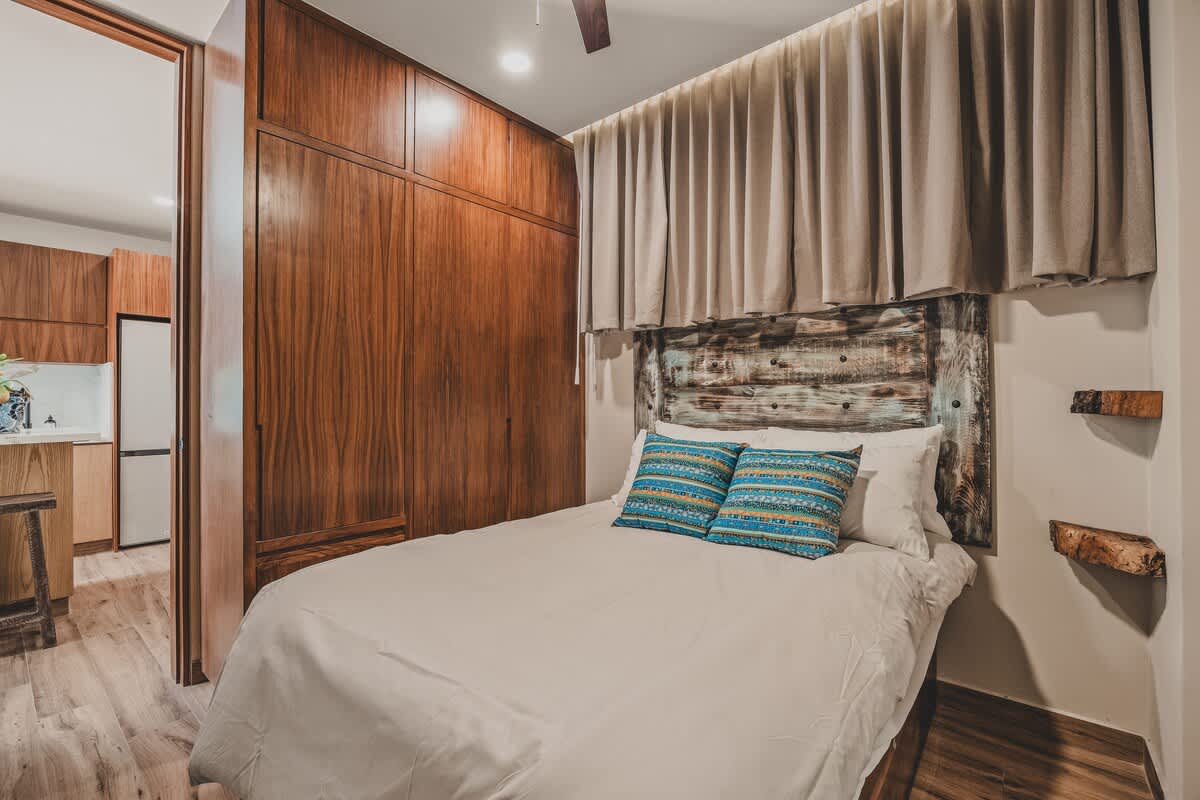 Bedroom with a bed featuring a distressed wooden headboard, light bedding, and blue patterned pillows. A large built-in wooden wardrobe spans the wall, and a ceiling fan is overhead.