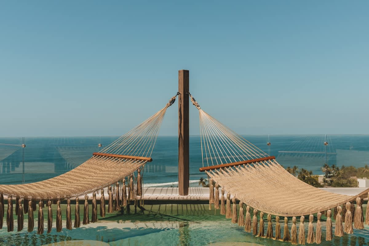 Two woven hammocks with decorative tassels are suspended over a sparkling infinity pool, offering a serene view of the vast blue ocean and distant horizon. The scene is bathed in bright sunlight, creating a relaxing outdoor ambiance.