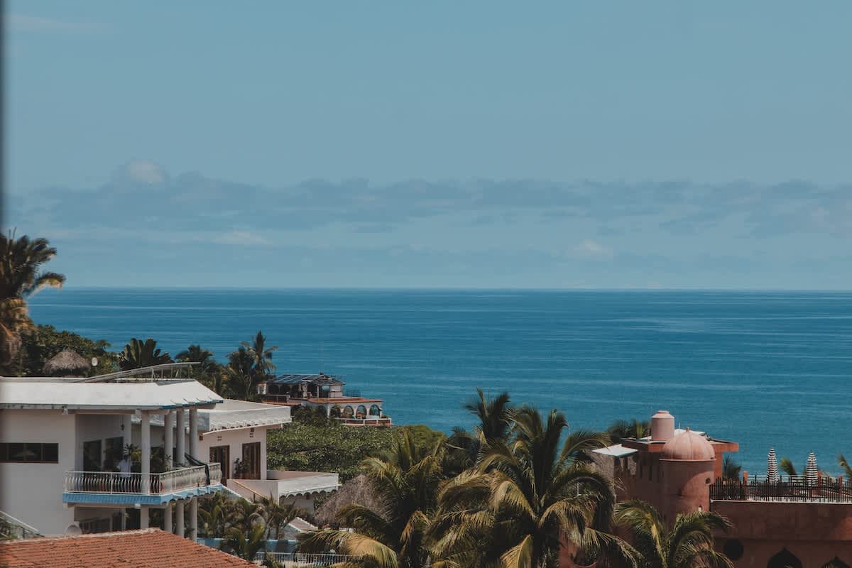 A panoramic view showcases several white and terracotta-colored buildings nestled among abundant palm trees, all overlooking the expansive blue ocean under a clear sky. The scene captures a vibrant coastal town or resort area.