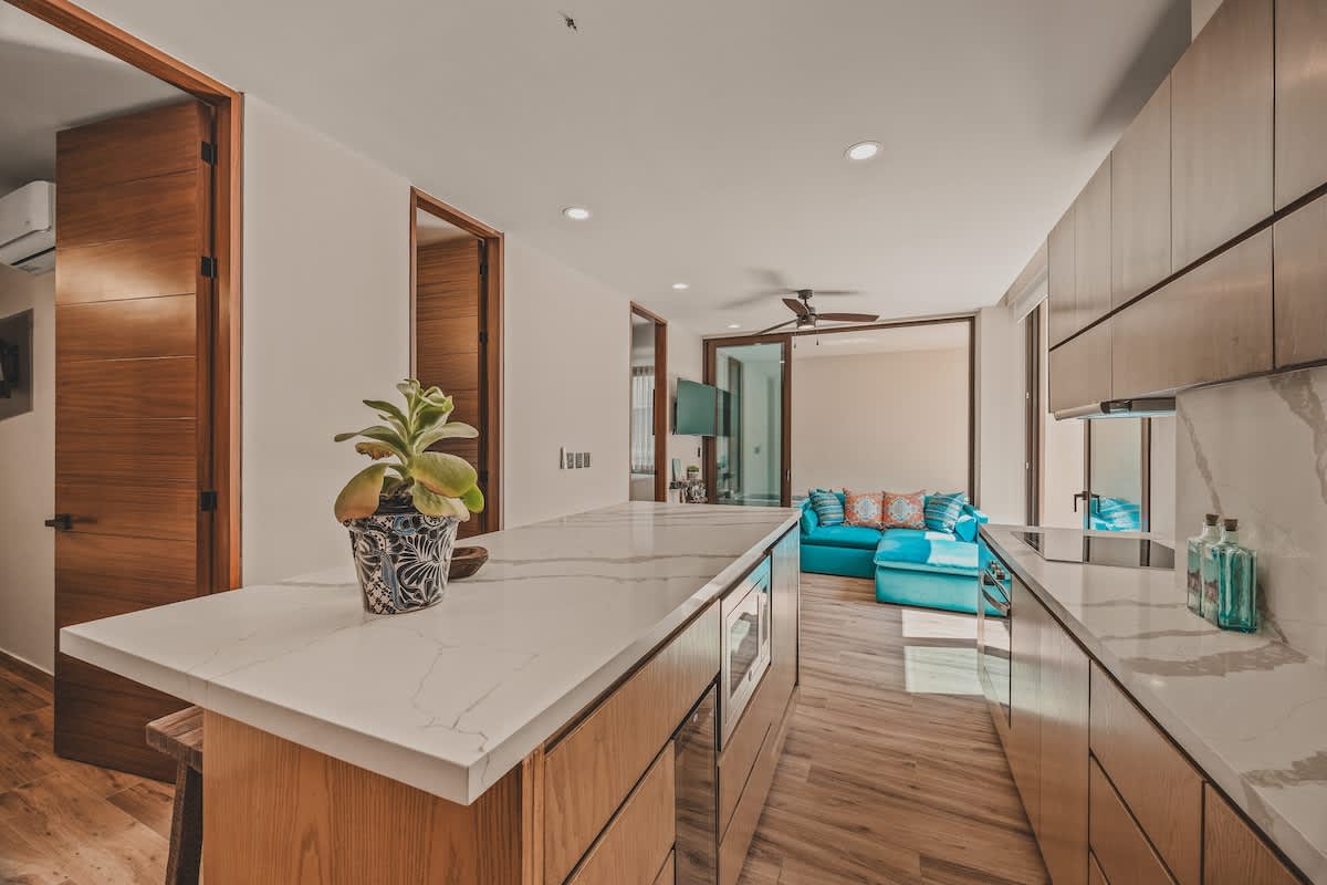 A sleek kitchen island with a white marble countertop and light wood base is the focal point, featuring built-in appliances and rustic wooden stools, with a view towards a modern bathroom entrance.