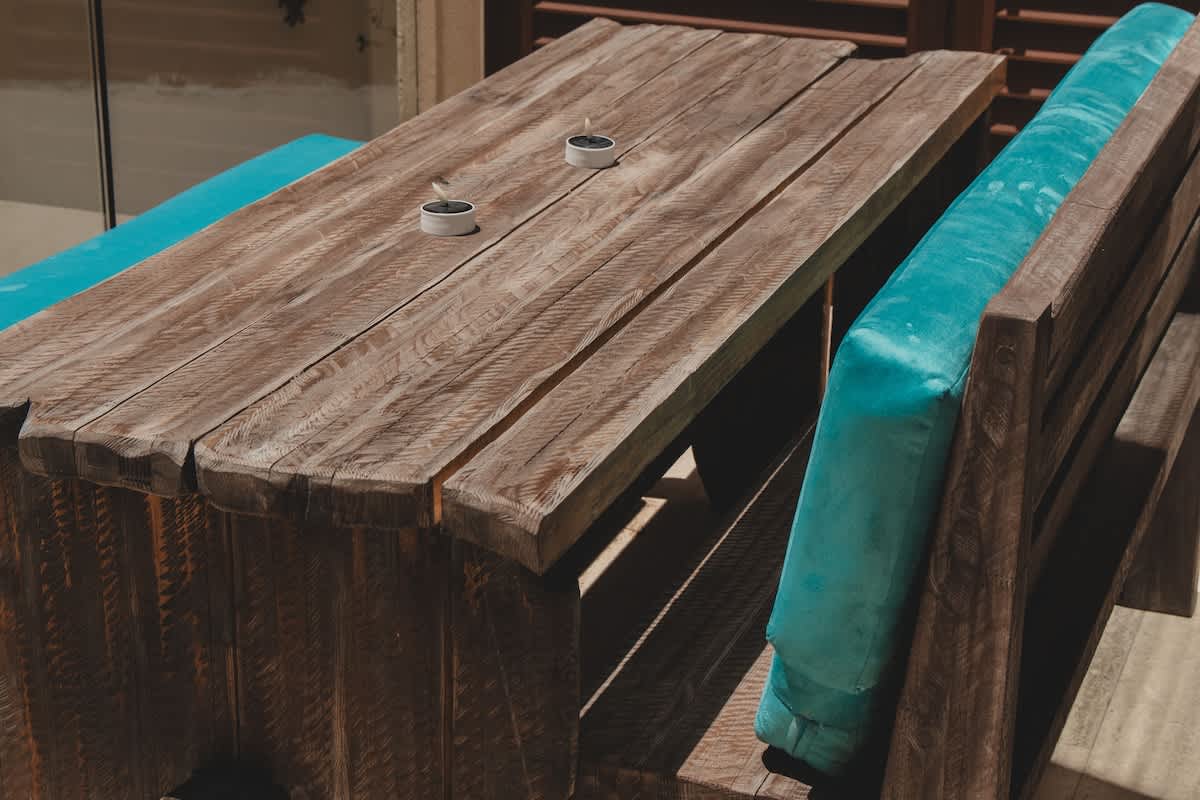 A sturdy, rustic wooden picnic table with benches featuring plush turquoise cushions is set outdoors, suggesting an al fresco dining or lounging spot. Two small white candle holders are placed on the table, ready for evening ambiance.