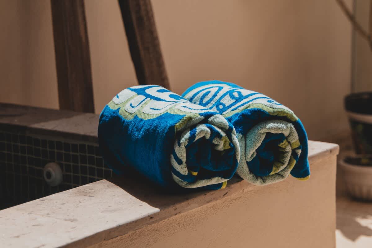 Two vibrant blue and green patterned towels are neatly rolled and placed on the light-colored concrete edge of an outdoor pool or hot tub, illuminated by natural sunlight.