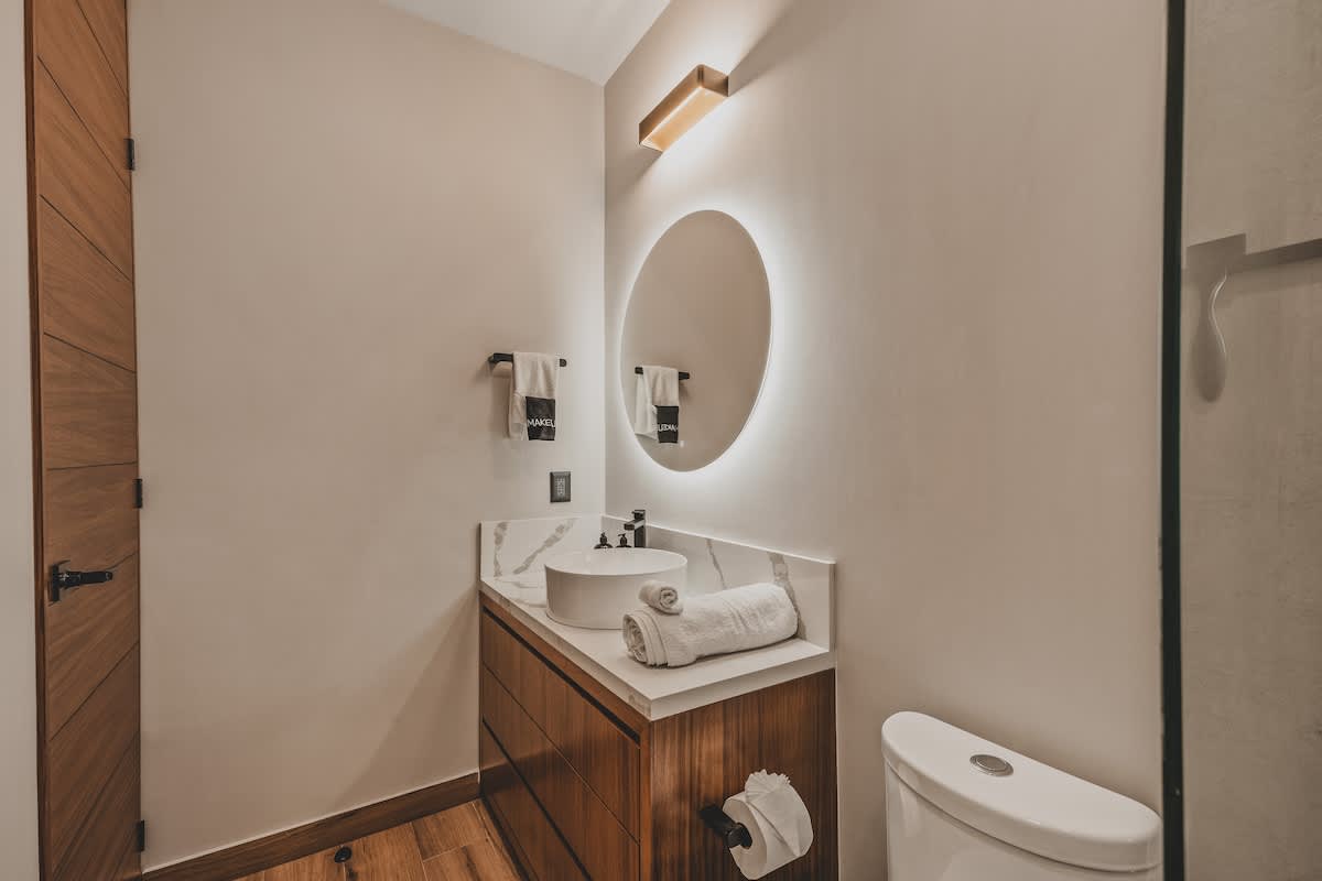Contemporary bathroom featuring a white vessel sink on a marble vanity, a round backlit mirror, and a wooden vanity cabinet. White towels are neatly folded on the counter, with a toilet visible to the right.