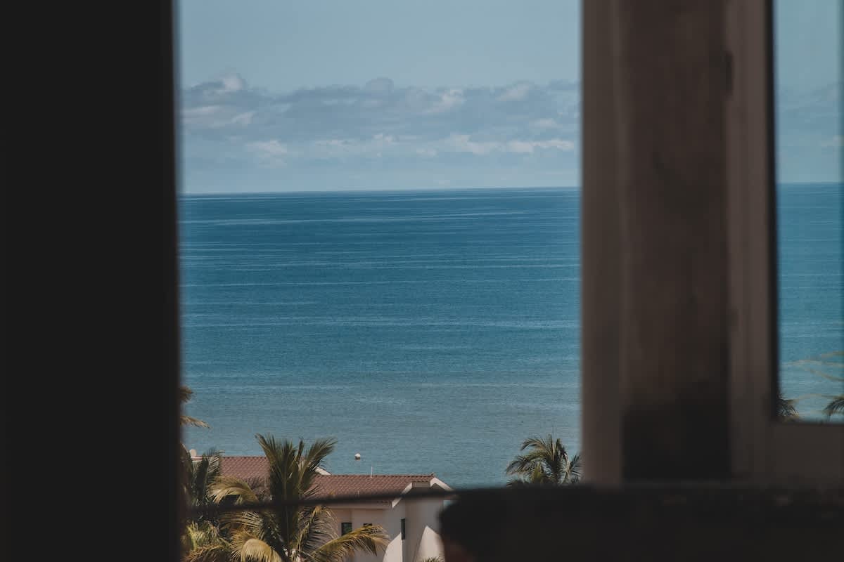 A framed view through a window reveals the vast, calm blue ocean under a partly cloudy sky. Below, the red-tiled roof of a building and lush palm trees are visible, suggesting a coastal property.