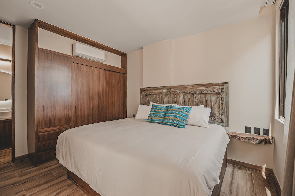 Spacious bedroom featuring a large bed with a rustic wooden headboard, light bedding, and two blue patterned pillows. A built-in wooden wardrobe with an AC unit is on the left, and a live-edge wooden nightstand is on the right.
