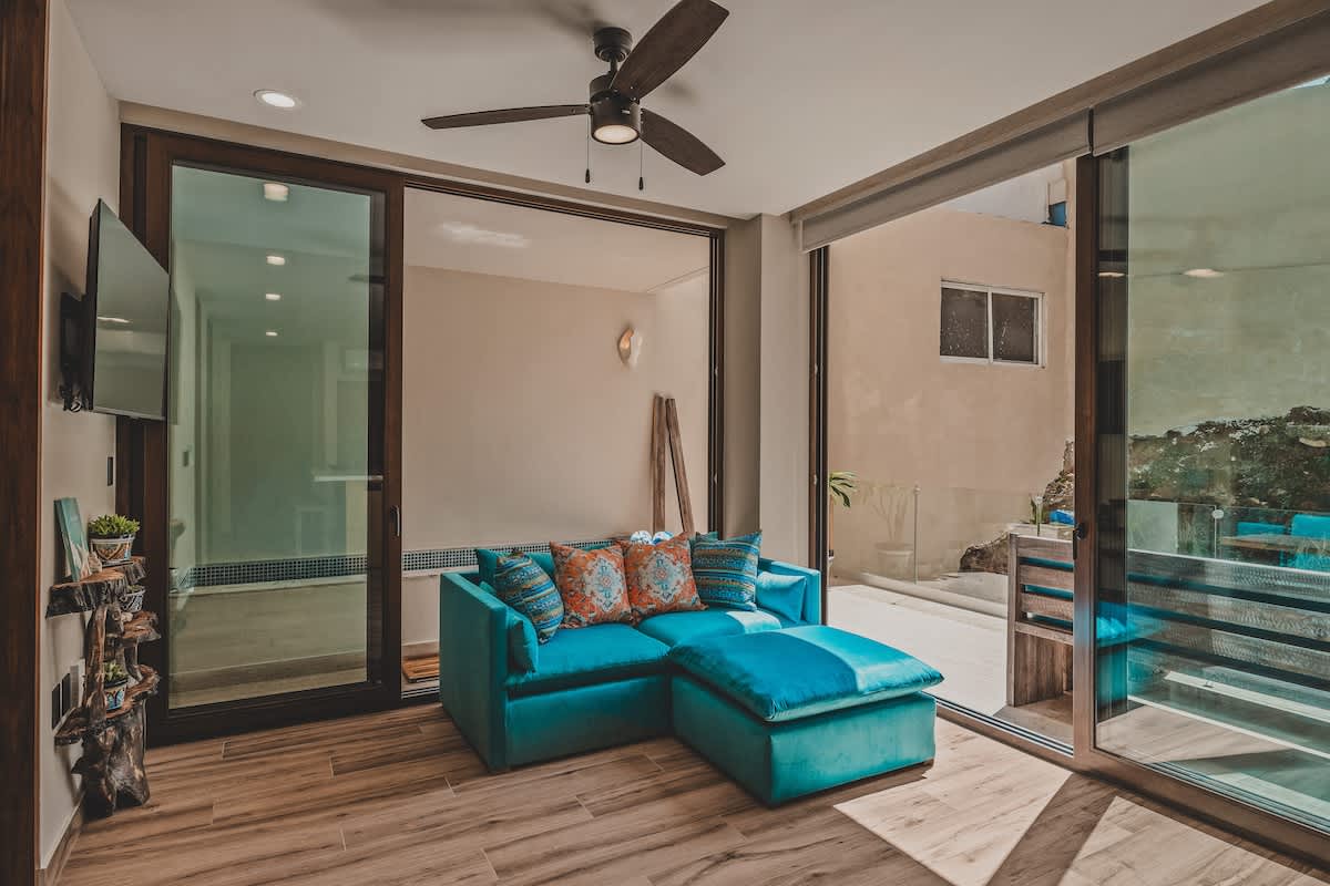 A cozy living room features a vibrant turquoise sectional sofa with decorative pillows, a ceiling fan, and large sliding glass doors that open to an outdoor patio with dining furniture.