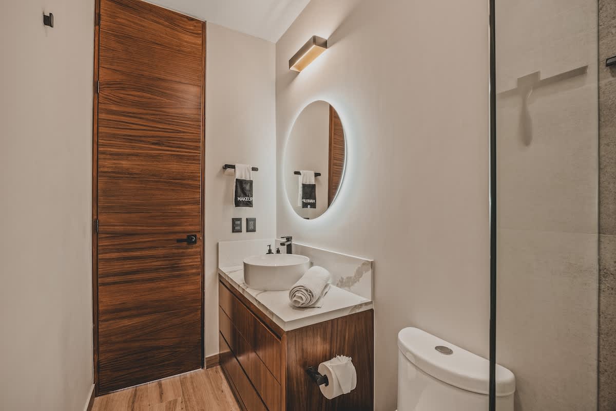 Contemporary bathroom with a sleek white vessel sink on a marble vanity, a round backlit mirror, and a toilet. A dark wooden door and light walls complete the clean aesthetic.