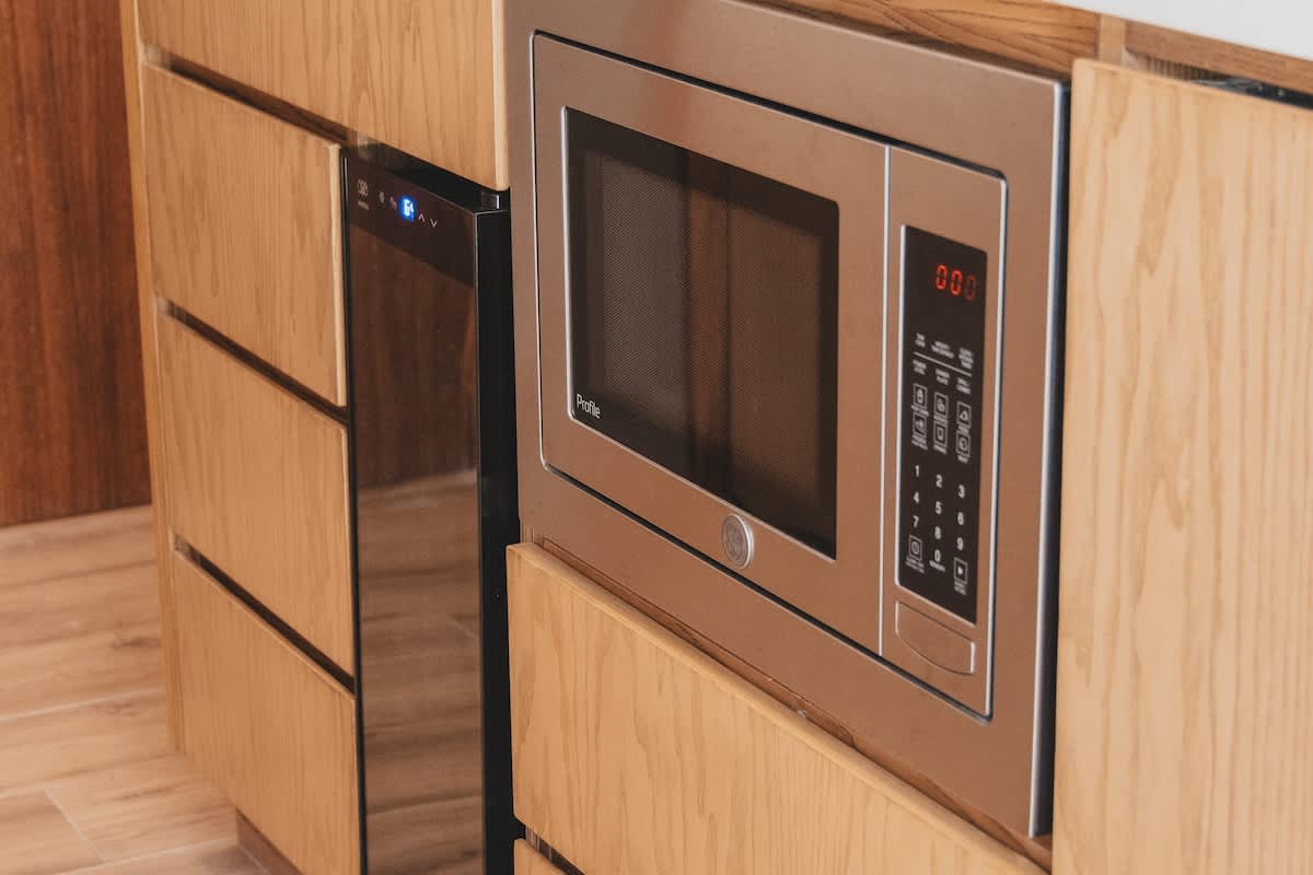 Built-in stainless steel microwave oven with digital display and control panel, integrated into light wood cabinetry, next to a compact beverage cooler.