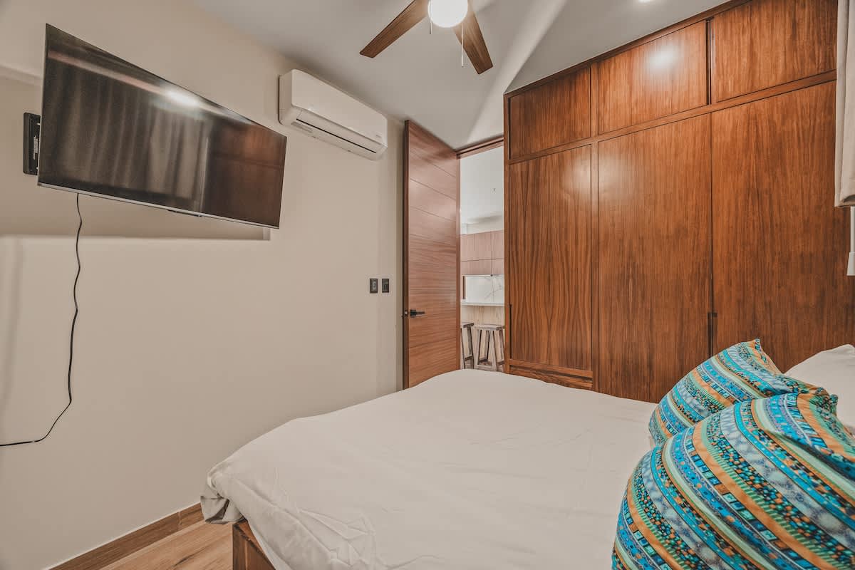 Bedroom featuring a wall-mounted TV, an air conditioning unit, and a large built-in wooden wardrobe. The bed has blue patterned pillows and light bedding, with a ceiling fan overhead.