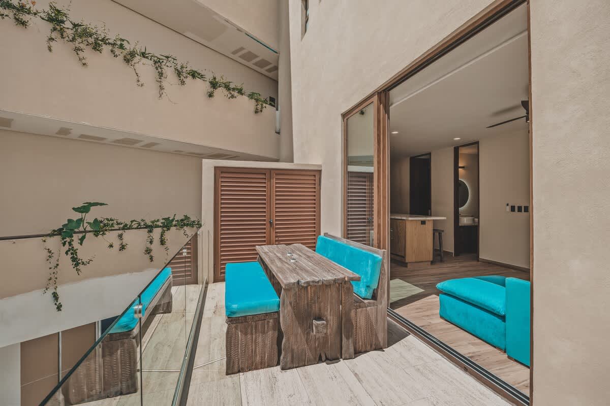 This elevated outdoor patio features a rustic wooden table with built-in benches and bright turquoise cushions, overlooking a lower level through a glass railing. A large sliding glass door opens to an interior space with a modern vanity, and light-colored walls are adorned with climbing greenery.