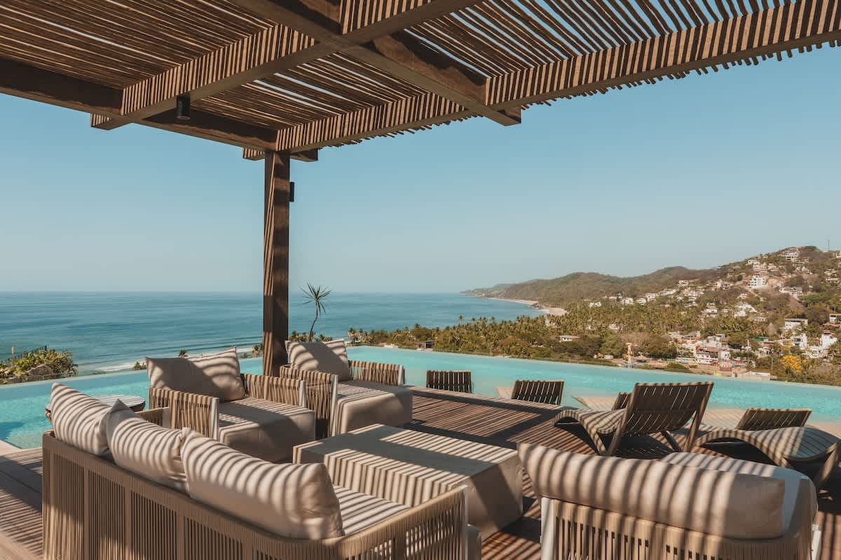 Expansive rooftop terrace featuring an infinity pool that blends seamlessly with the ocean horizon, surrounded by comfortable outdoor lounge seating under a slatted wooden pergola. Offers breathtaking coastal views.