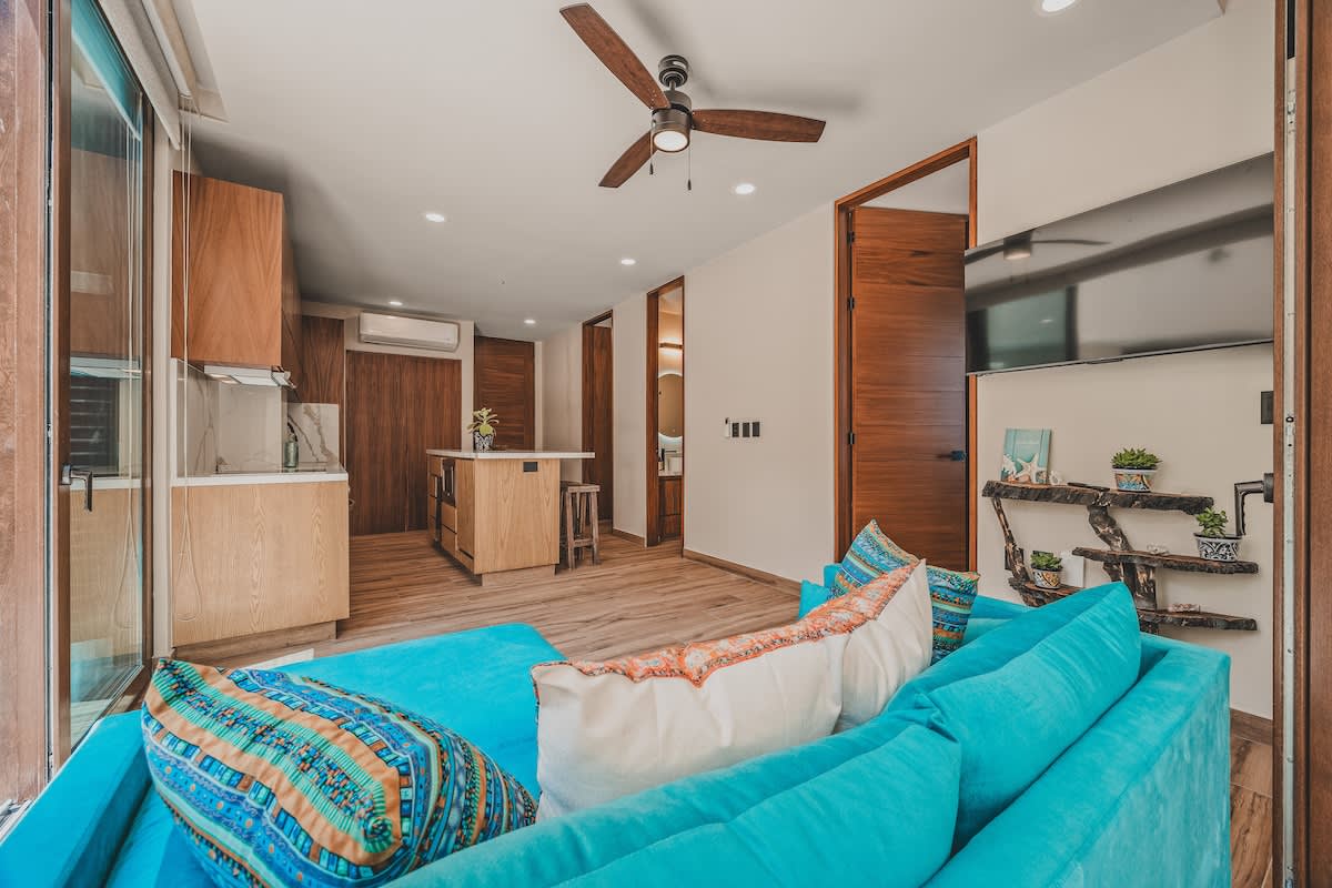Bright open-concept living space featuring a vibrant blue sofa with patterned pillows, a wooden kitchen island with bar stools, and light wood flooring.
