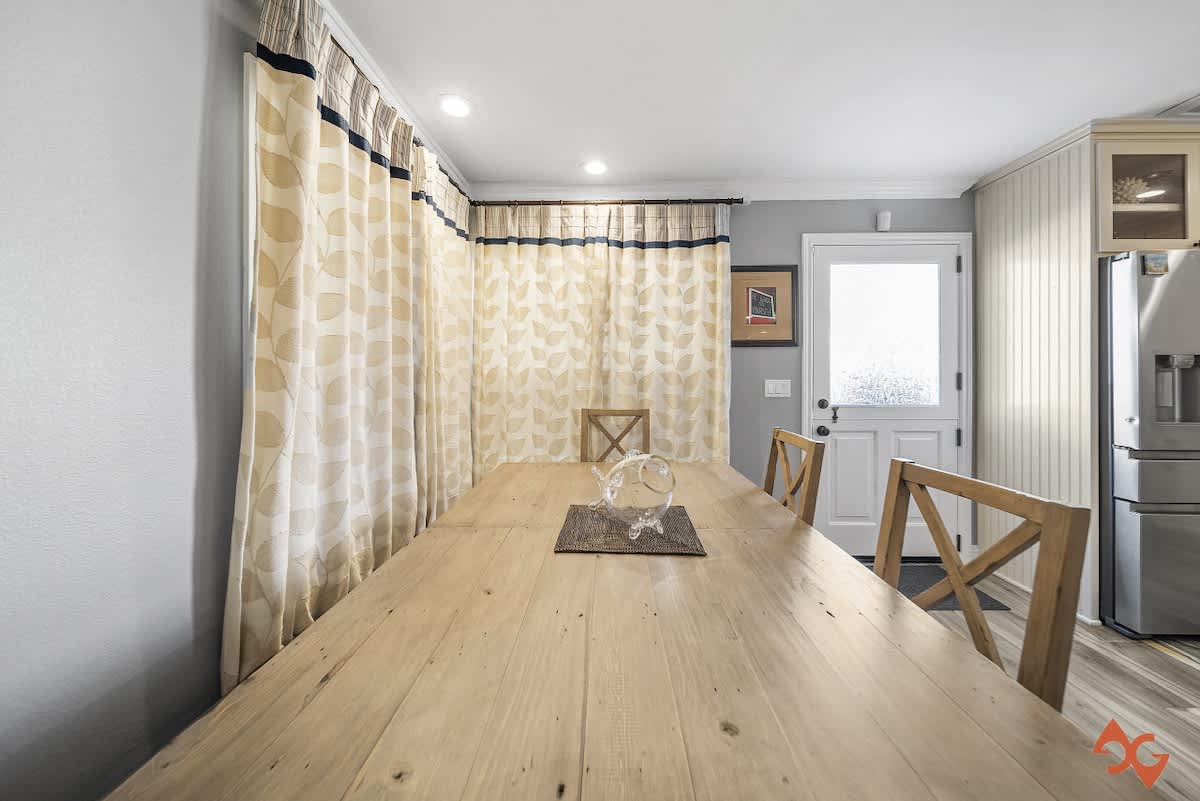 A bright dining room features a long, rustic wooden table with cross-back chairs, light-colored patterned curtains, and wood-look flooring, with a glimpse of a stainless steel refrigerator in the background.