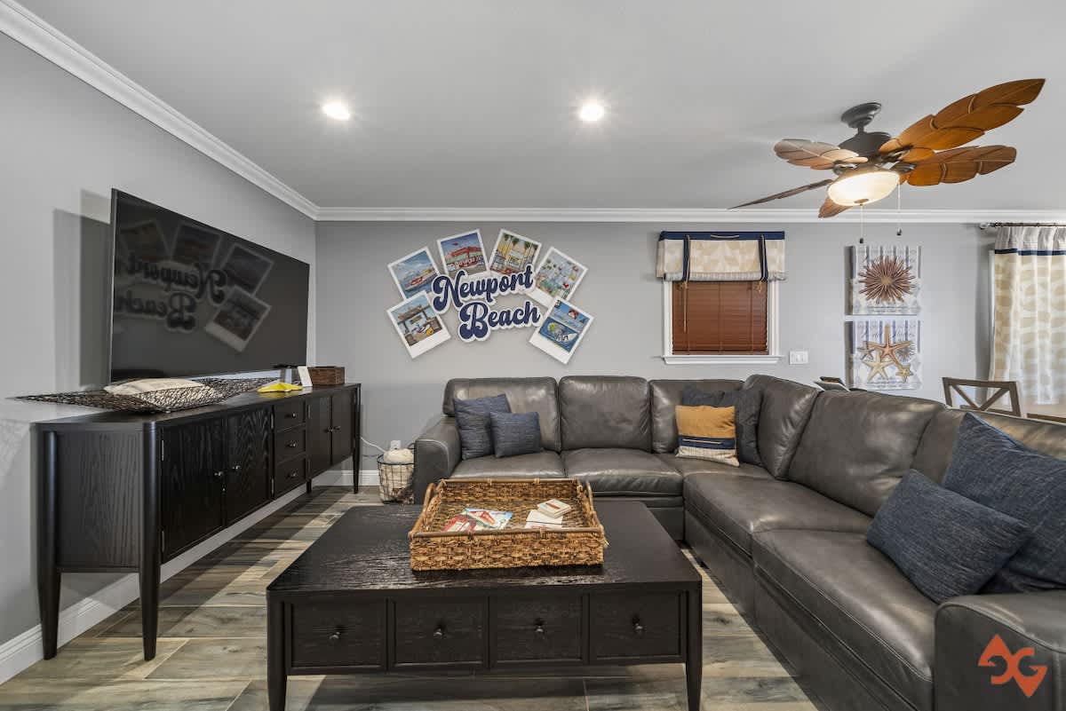 A spacious living room featuring a large gray sectional sofa, a dark wood coffee table with a woven basket, and a wall-mounted TV. Nautical-themed decor, including a "Newport Beach" sign and starfish art, adorns the light gray walls.