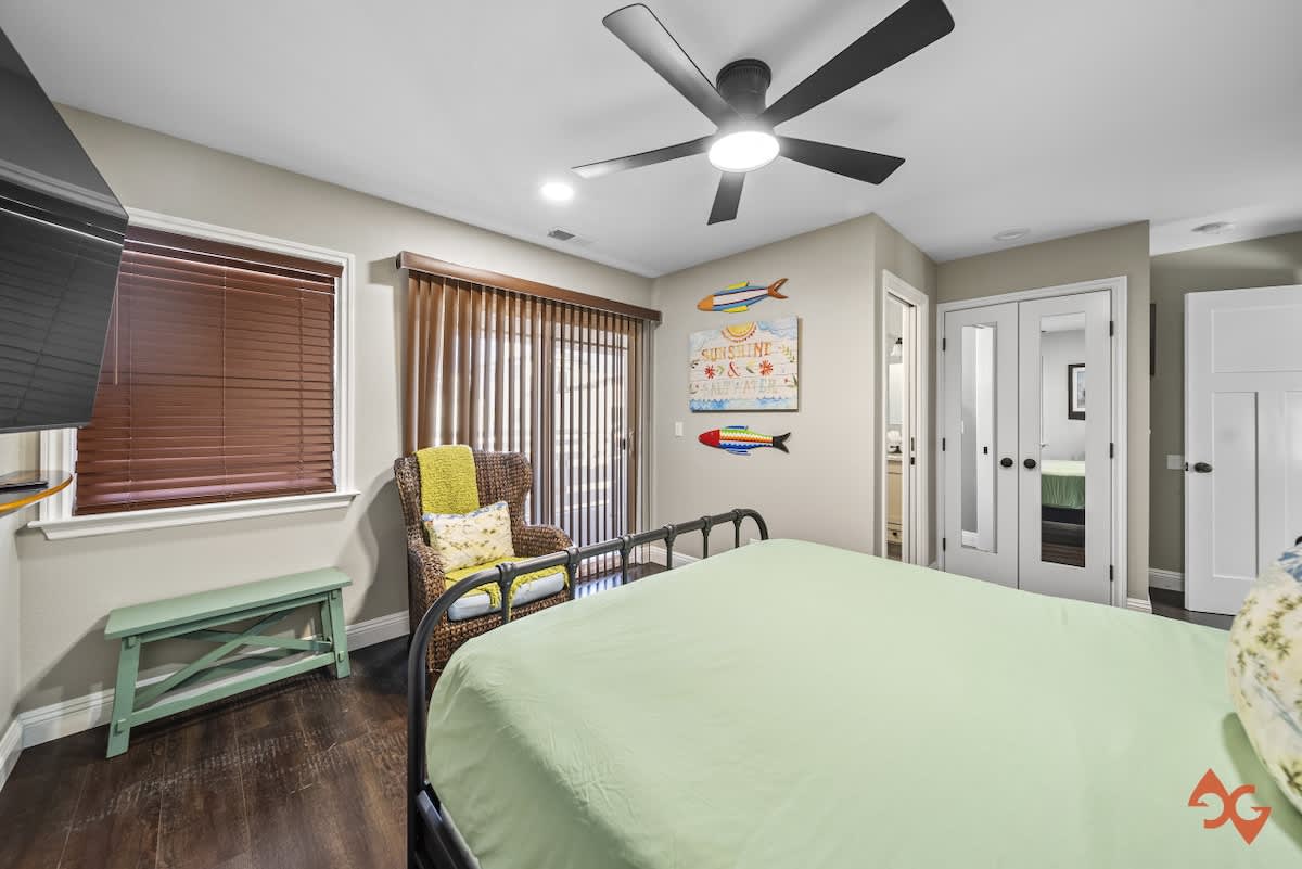 A bedroom with a black metal bed frame, a wicker armchair with a yellow throw, and coastal wall art, featuring a sliding glass door with vertical blinds leading to an outdoor area.