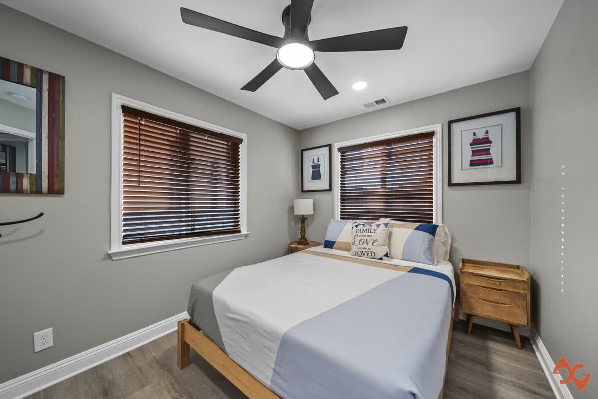 A bright bedroom with light gray walls, a queen-sized bed with blue and white bedding, and two windows with dark wood blinds. A black ceiling fan with a light fixture is centered, and framed artwork of swimsuits hangs on the walls.
