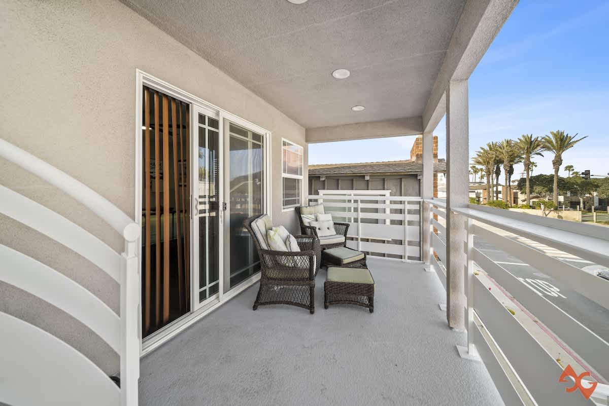 A covered outdoor balcony features wicker armchairs with light cushions and an ottoman, set against a white slatted railing and a view of palm trees and a street. Sliding glass doors lead into the interior.